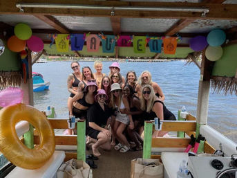 Group of friends in swimsuits posing on a decorated, covered party boat at a sunny waterfront with colorful balloons, a festive banner and an inflatable life ring.