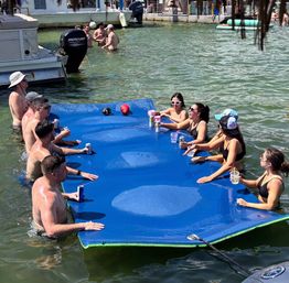 Sunny lakeside summer scene of adults in swimsuits socializing around a large blue floating mat with drinks, anchored near boats and a dock in green water at a busy marina.