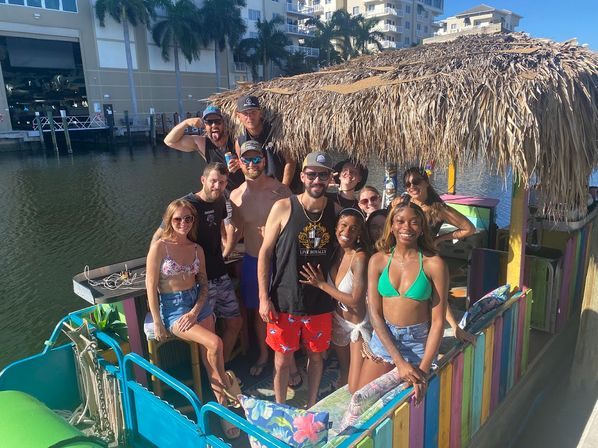 Group of friends on a colorful tiki-style pontoon boat at a tropical marina, wearing swimsuits and sunglasses, smiling under a thatched palapa with palm trees and waterfront buildings in the sunny dockside background.