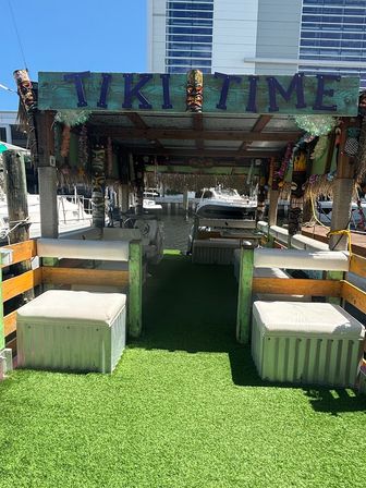 Sunny waterfront tiki dock lounge at a coastal marina with a 'TIKI TIME' sign, boats, cushioned benches and artificial grass