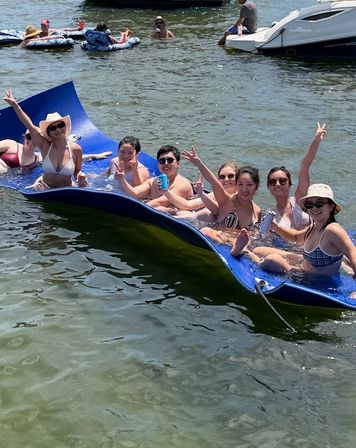 Group of friends enjoying a sunny lake day on a large blue floating mat, waving and holding drinks near boats and inner tubes.