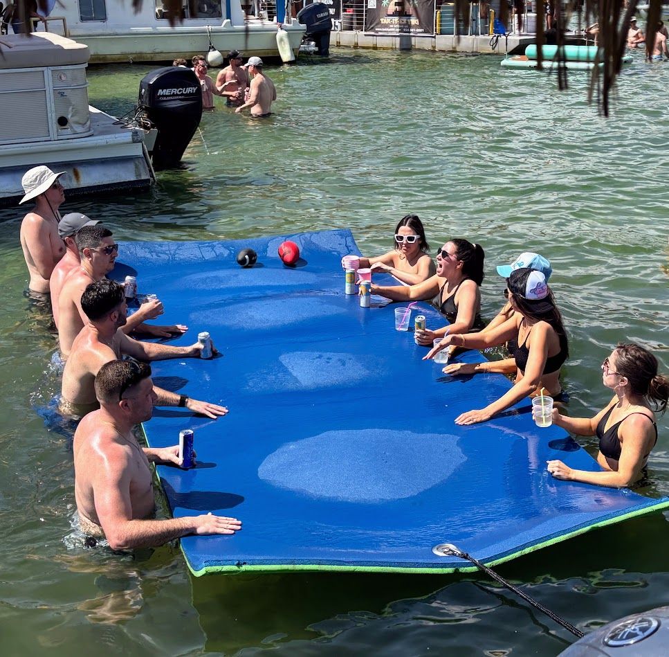 Group of friends lounging around a large blue floating mat in shallow lake water by a boat dock, holding drinks and socializing at a sunny summer lake party.