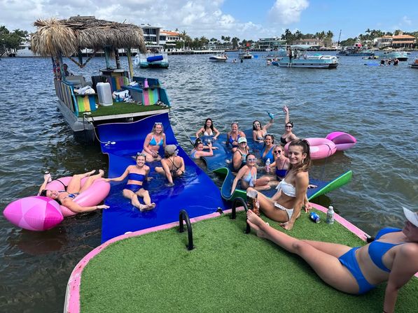 Group of friends in swimwear at a sunny boat party on a coastal waterway — tiki-roofed floating bar, blue inflatable slide, pink floats and anchored boats near waterfront homes.