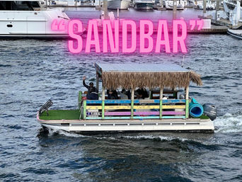 Colorful tiki-style pontoon boat with thatched roof and turf deck carrying waving passengers on a marina waterway, pink "SANDBAR" sign overlay and yachts in the background.