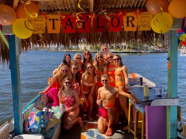 Cheerful group in swimsuits posing on a colorful tiki-style party boat at a sunny waterfront, decked with balloons, tropical cushions and a festive banner.