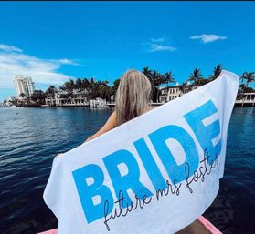 Person on a boat draped in a white towel that reads "BRIDE", overlooking a palm-lined tropical waterfront with blue sky and luxury homes