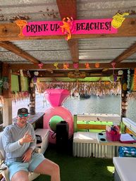 Waterside tiki bar with bright pink "DRINK UP BEACHES!" sign, straw-thatched roof, colorful tropical decorations, a large pink inflatable ring, and a person in sunglasses flashing a shaka hand gesture.