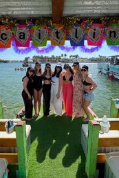 Seven friends in swimsuits and hats posing on a decorated pontoon dock under a colorful banner, turquoise water, palm trees and waterfront homes in the sunny coastal background.