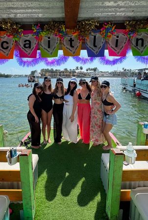 Seven friends in swimsuits and hats posing on a decorated pontoon dock under a colorful banner, turquoise water, palm trees and waterfront homes in the sunny coastal background.