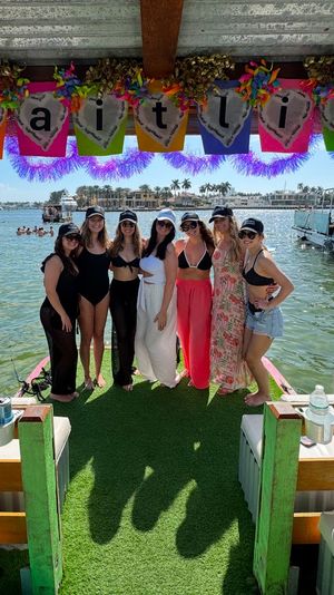 Seven friends in swimsuits and hats posing on a decorated pontoon dock under a colorful banner, turquoise water, palm trees and waterfront homes in the sunny coastal background.