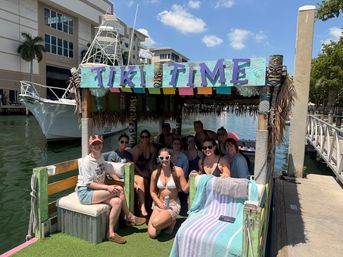 Smiling group of friends on a colorful "TIKI TIME" floating tiki bar at a sunny marina dock, tropical decor, towels and drinks for a fun boat party vibe.