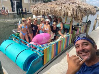 Smiling man makes a shaka in a selfie at a marina beside a colorful tiki-style pontoon boat; a group in swimsuits and straw hats pose under a thatched roof with inflatables and foam mats.