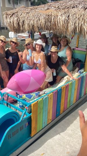 Smiling man makes a shaka in a selfie at a marina beside a colorful tiki-style pontoon boat; a group in swimsuits and straw hats pose under a thatched roof with inflatables and foam mats.
