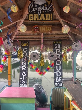 Colorful waterfront tiki bar on a docked boat decorated for graduation — thatched roof, 'Congrats Grad' banners, rainbow leis and garlands, gold tassels, paper lanterns, outboard motor, and marina reflections.