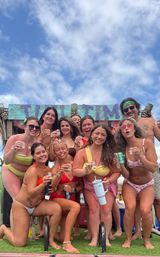 Cheerful group of friends in colorful bikinis holding drinks at a sunny tropical beach tiki-bar party under a blue sky