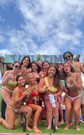 Cheerful group of friends in colorful bikinis holding drinks at a sunny tropical beach tiki-bar party under a blue sky
