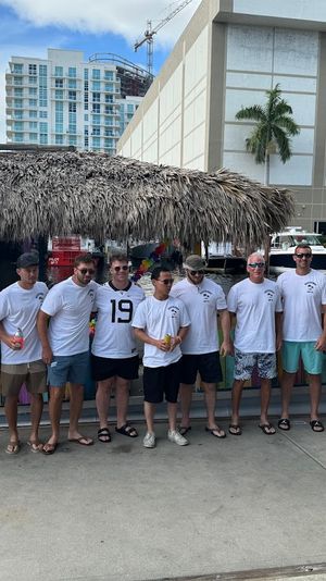 Group of men in matching white shirts standing on a marina dock in front of a colorful tiki-style boat with a thatched roof, palm trees and waterfront high-rise buildings