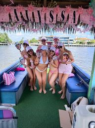 Group of women in bikinis and matching hats posing on a pink-decorated party boat along a sunny waterfront canal — playful bachelorette/boat party vibe.
