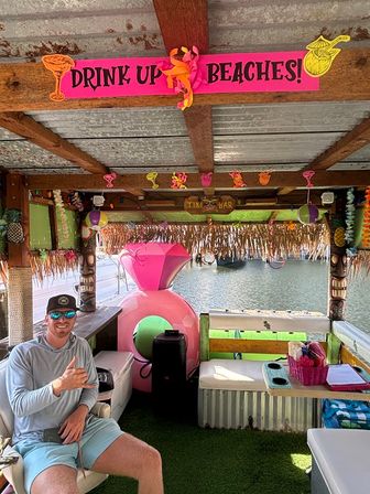 Bright dockside tiki bar with a pink 'DRINK UP BEACHES!' sign, tropical decorations, a large pink inflatable float, cushioned seating and a person in sunglasses and cap flashing a shaka gesture, overlooking calm lake water.