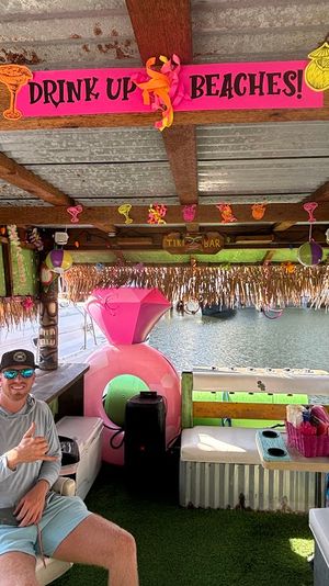 Bright dockside tiki bar with a pink 'DRINK UP BEACHES!' sign, tropical decorations, a large pink inflatable float, cushioned seating and a person in sunglasses and cap flashing a shaka gesture, overlooking calm lake water.