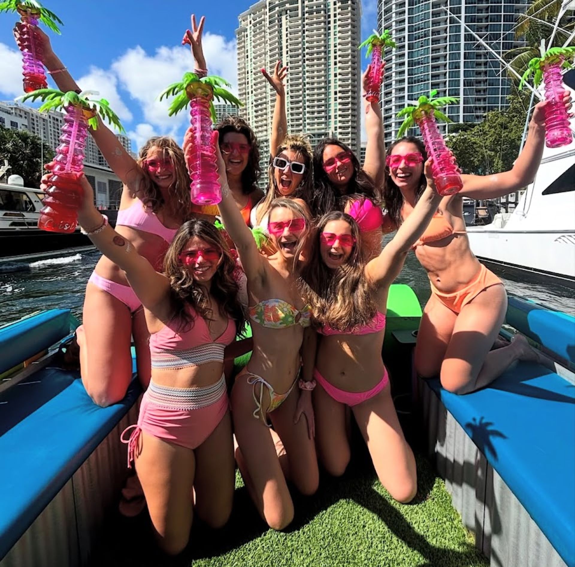 Group of friends in colorful bikinis and pink sunglasses cheering on a boat party at a sunny waterfront marina, holding tall pink palm-shaped drink cups with high-rise coastal condos and palm trees in the background.