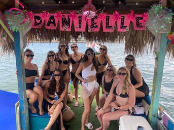 Group of women in bikinis and sunglasses posing under a thatched tiki hut over water, holding drinks beneath pink party decorations and a small disco ball on a floating dock.