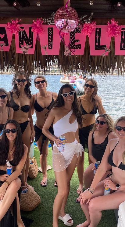 Group of women in bikinis and sunglasses posing under a thatched tiki hut over water, holding drinks beneath pink party decorations and a small disco ball on a floating dock.
