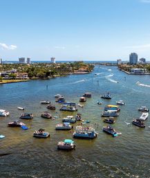 Aerial view of a sunny coastal waterway dotted with dozens of anchored pontoons and motorboats hosting a lively boat party near waterfront homes and a distant high-rise skyline.