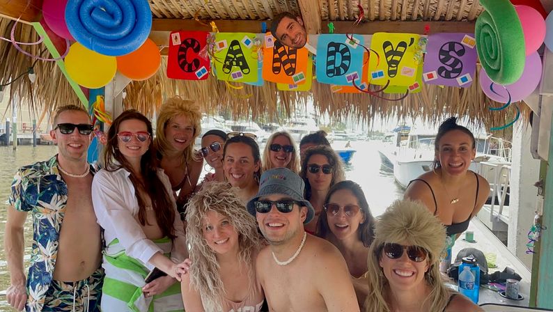 Group of friends smiling and posing under a decorated thatched tiki hut at a dockside birthday boat party, colorful balloons and banner overhead with marina and boats in the background.
