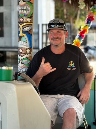 Smiling man in a cap and sunglasses seated at a boat helm, wearing a tiki-style shirt and flashing a shaka sign beside a colorful carved tiki pole and lei garland at a sunny marina.
