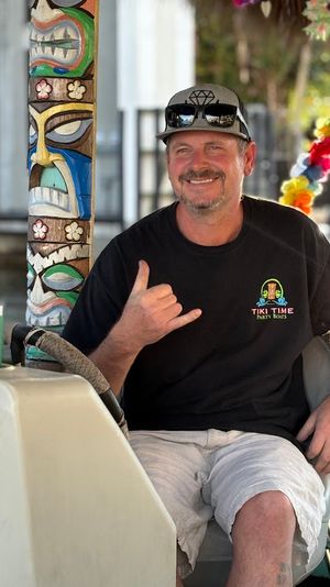 Smiling man in a cap and sunglasses seated at a boat helm, wearing a tiki-style shirt and flashing a shaka sign beside a colorful carved tiki pole and lei garland at a sunny marina.