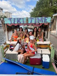 Smiling group of women on a decorated tiki-style party boat along a sunny canal, bachelorette sash and champagne, coolers and surfboard, waterfront buildings and trees in background.