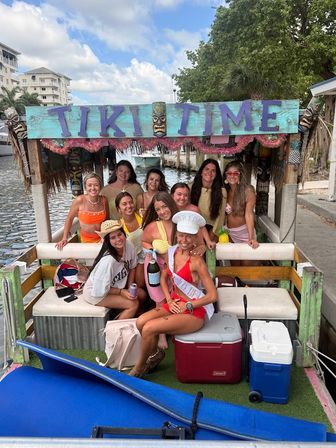 Smiling group of women on a decorated tiki-style party boat along a sunny canal, bachelorette sash and champagne, coolers and surfboard, waterfront buildings and trees in background.