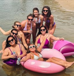 Group of women in purple and pink swimsuits wearing heart-shaped sunglasses, smiling and holding canned drinks while lounging on a pink inflatable float in shallow water at a sandy lakeshore.