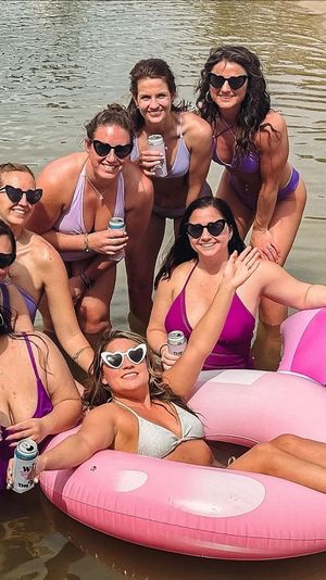 Group of women in purple and pink swimsuits wearing heart-shaped sunglasses, smiling and holding canned drinks while lounging on a pink inflatable float in shallow water at a sandy lakeshore.