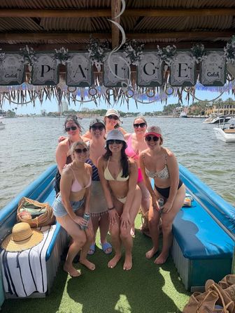 Seven friends in bikinis posing on a decorated tiki-style party boat, smiling on a sunny coastal marina with palm trees and docked yachts.