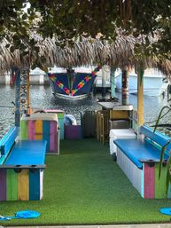Colorful tiki-hut dock at a tropical marina with a thatched roof, rainbow-painted benches and blue cushions, artificial grass floor and a leis-decorated boat moored on the water.
