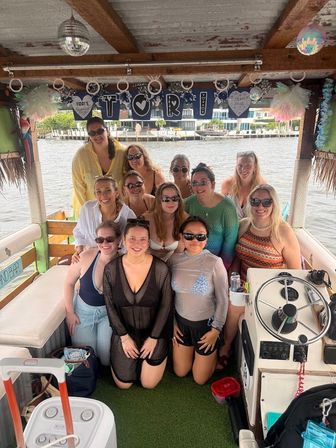 Smiling group of women in swimsuits and sunglasses posing on a decorated party boat at a marina with waterfront homes visible in the background.