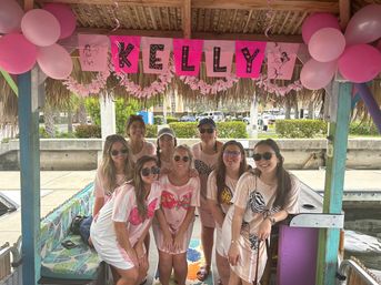 Group of women in matching pink shirts posing under a decorated tiki hut at a marina, with pink balloons, a festive name banner overhead, and dock and palm trees in the background.