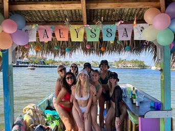 Group of women in swimsuits smiling inside a tiki-style floating cabana on a sunny bay, pastel balloons and a colorful name banner overhead, drinks on the bar and waterfront homes in the background.