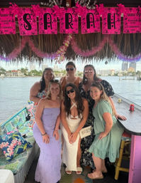 Six women, including a bride in a white dress with a 'bride' bag, posing on a decorated waterfront tiki-style boat bar under pink bachelorette banners and leis with a city skyline across the water