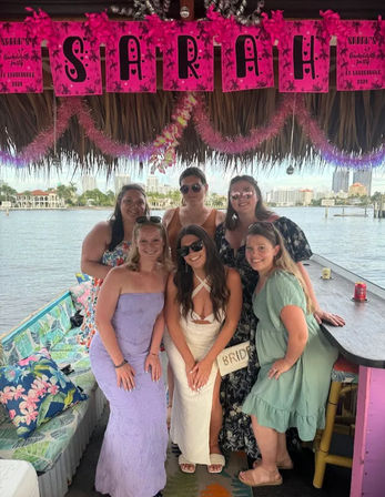 Six women, including a bride in a white dress with a 'bride' bag, posing on a decorated waterfront tiki-style boat bar under pink bachelorette banners and leis with a city skyline across the water