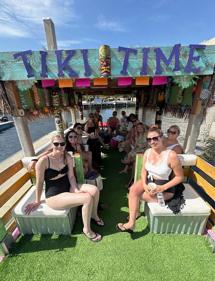 Group of friends on a colorful tiki-themed party boat docked at a marina, sitting under a 'TIKI TIME' sign in swimsuits and sunglasses on a sunny waterfront day.
