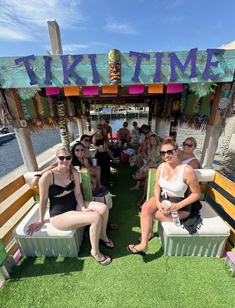 Group of friends on a colorful tiki-themed party boat docked at a marina, sitting under a 'TIKI TIME' sign in swimsuits and sunglasses on a sunny waterfront day.