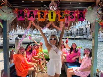 Group of women on a tiki-style boat at a marina celebrating a bachelorette — bride in a white sash raises her arms while friends in pink and orange hold tropical drinks, with waterfront docks and condos in the background.