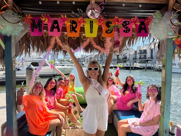 Group of women on a tiki-style boat at a marina celebrating a bachelorette â bride in a white sash raises her arms while friends in pink and orange hold tropical drinks, with waterfront docks and condos in the background.