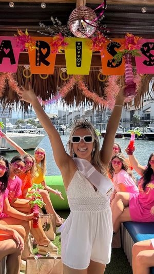 Group of women on a tiki-style boat at a marina celebrating a bachelorette — bride in a white sash raises her arms while friends in pink and orange hold tropical drinks, with waterfront docks and condos in the background.