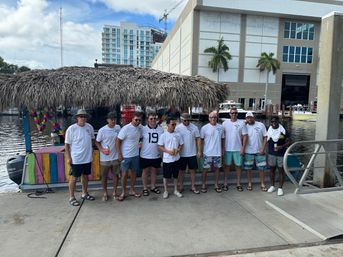 Eleven men in matching white shirts posing on a waterfront dock beside a colorful tiki boat with a thatched roof, palm trees, boats and a high-rise marina in the background.
