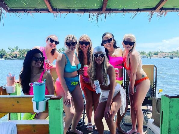 Seven women in colorful bikinis and sunglasses posing on a covered boat by a palm-lined waterfront, smiling and holding drinks on a sunny day.