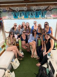 Group of smiling women on a decorated daytime party boat with waterfront homes in the background, personalized banner overhead, tote bags and party props, one visibly pregnant guest.